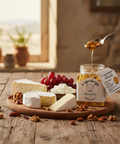 Honey being drizzled from a spoon over a jar of honey with cheese and grapes on a wooden board.