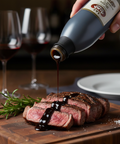 Sliced steak on a wooden board with a bottle of soy sauce being poured over it, set against a backdrop of wine glasses.