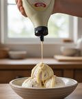 Hand pouring a creamy sauce from a bottle labeled 'Kewpie' onto ice cream in a bowl.