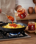 Person adding tomato sauce from a jar to pasta in a pan on a stove.