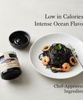 Black squid ink pasta with prawns on a white plate, next to a jar of black squid ink, on a light gray background.