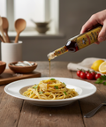 Person pouring olive oil over spaghetti on a wooden table with kitchen utensils and ingredients in the background.
