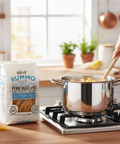 Person cooking gluten-free pasta in a pot with a package of Rummo pasta on a kitchen counter.