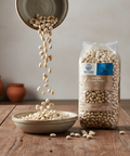White beans being poured from a bowl into another, with a Del Colle branded package of beans on a wooden surface.