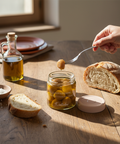 Person dipping bread into a jar of olive oil on a wooden table with a window in the background.