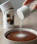 Hand sprinkling sugar into a bowl of chocolate dessert with a container of sugar in the background.