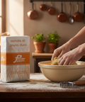 Person kneading dough in a bowl with a box of Fainimo Pasini semolina on a wooden table.