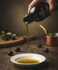Hand pouring olive oil from a bottle into a white dish on a wooden surface with olives in the background.