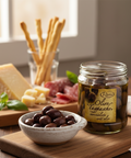 Jar of olives on a wooden board with a bowl of olives and charcuterie in the background.