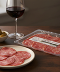 Plated salami slices with a glass of red wine and olives on a wooden table.