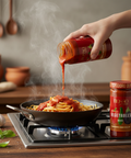 Person pouring sauce from a jar onto pasta in a pan on a stove, with additional jars of sauce in the background.