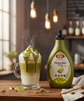 Pistachio sauce bottle next to a pistachio latte on a wooden table with a blurred background.
