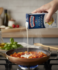 Person pouring cream from a carton into a pan of pasta on a stove