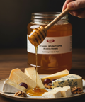Honey being drizzled from a wooden dipper onto a plate of cheese and grapes with a jar of honey in the background.